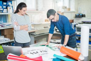 Confident young specialists standing at table full of various types of paper and discussing color palette while going to print banner in advertising company office